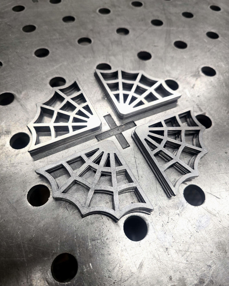 Two 5-inch spider web decorative steel gussets placed on a metal fabrication welding table with circular mounting holes.