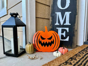 A bright orange steel jack-o'-lantern with a wide toothy grin placed on a front porch next to a "Welcome" leaning sign and a black decorative lantern.