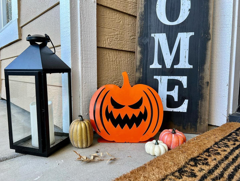 A bright orange steel jack-o'-lantern with a wide toothy grin placed on a front porch next to a "Welcome" leaning sign and a black decorative lantern.